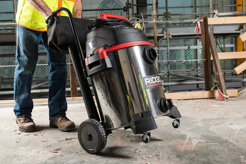 16-Gallon Stainless Steel Wet Dry Shop Vacuum With Cart in use on a construction site, showing large rear wheels and push handle.