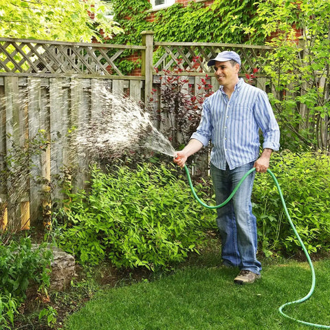 Person watering a garden with a green hose next to a wooden fence, wearing jeans and a striped shirt, surrounded by lush greenery.