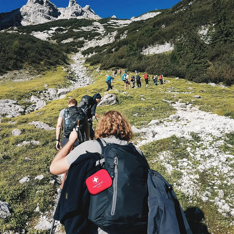Hikers ascending a mountain trail with a visible Mini First Aid Kit 120 Piece Waterproof Hard Shell Medical Kit For Car Home Office Travel Camping Sports Outdoor School Emergency Supplies And Survival Gear Red.