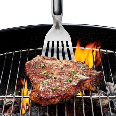 Grilling Tool Precision Turner flipping steak over grill with thin beveled head, non-slip grip, and hanging ring, OXO Good Grips Black