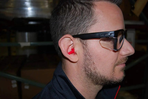 Man wearing ANSI Z87.1 Safety Glasses With Anti Fog UV Protection Clear Lens Side Shields indoors, showing side view with red earplugs.