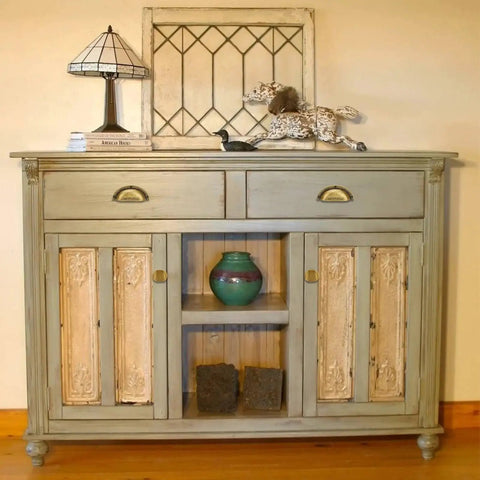 Antique wooden cabinet with two drawers and intricate detailing, decor items on top, featuring brass handles for drawers and doors.