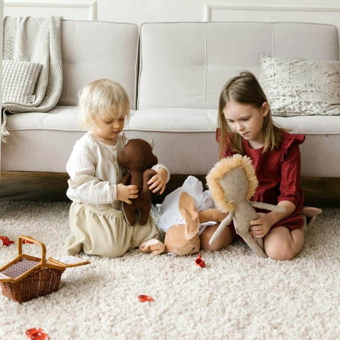 Two children playing with stuffed animals on a beige textured rug in a cozy living room setting.