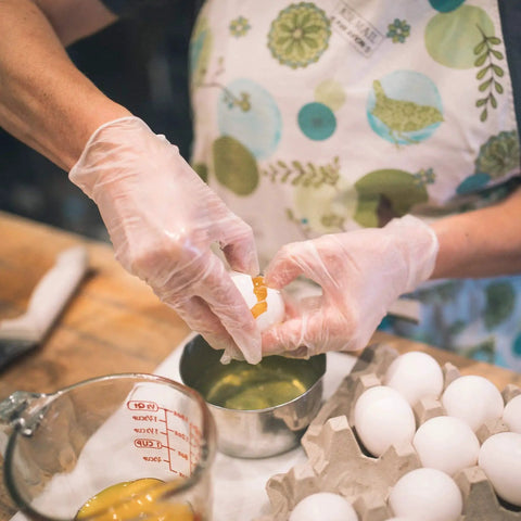 Person wearing Clear Vinyl Gloves Latex Free Powderfree Food Safe Disposable Nonsterile cracking an egg into a bowl