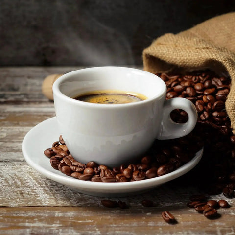 White coffee cup filled with hot espresso, surrounded by roasted coffee beans on a wooden surface with a burlap sack in the background.