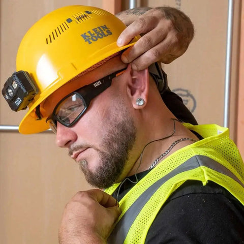Construction worker wearing ear protection, hard hat, and safety goggles on site, adjusting earplugs with hand.