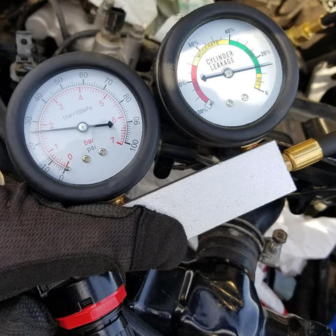 Automotive cylinder leak-down tester kit with dual gauges and extension rod, held by a gloved hand.