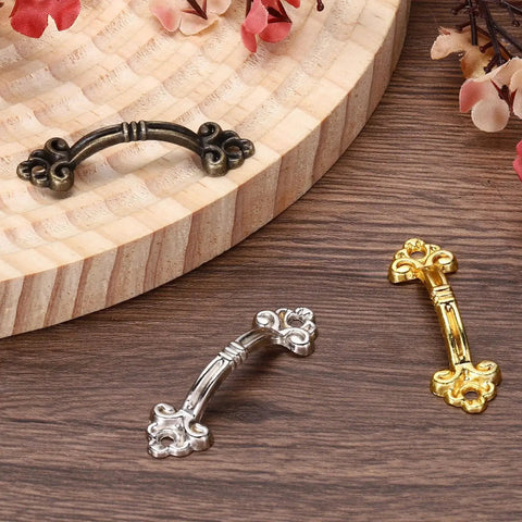 Three mini decorative drawer pulls in black, silver, and gold finishes on a wooden surface, showcasing the 20Pcs Mini Decorative Drawer Pulls Gold Finish 1.9 Inch Zinc Alloy Cabinet Handles With Screws For Furniture Dresser Jewelry Boxes And Sliding Doors Vintage Style Screw Mounted Hardware.