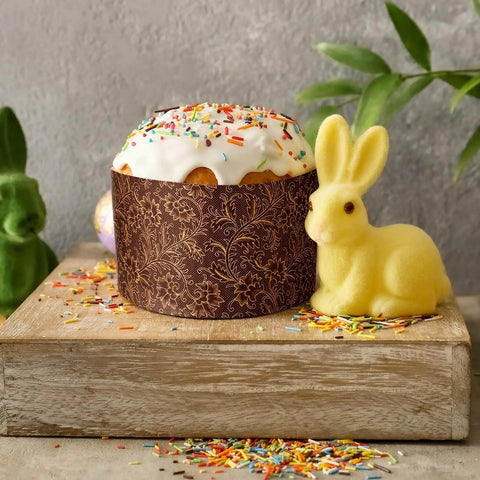Easter bread with frosting in a brown panettone paper mold, surrounded by a yellow bunny and colorful sprinkles on a wooden board.