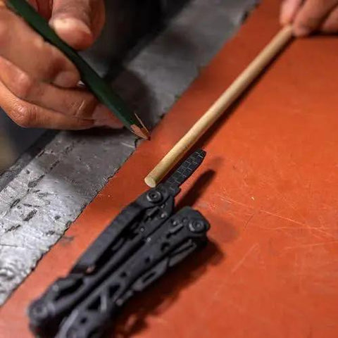Person using Suspension-NXT 15-In-1 EDC Multi Tool with pocket knife and pliers, next to a pencil and wooden dowel on a workbench.
