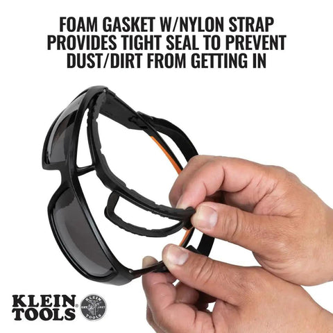 Hands holding full frame gasket safety glasses with foam seal and nylon strap, anti fog UV scratch resistant features.