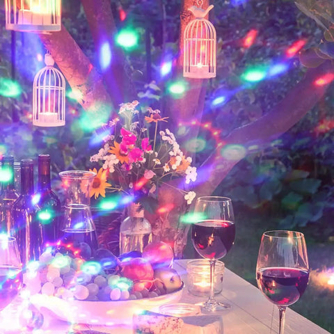 Romantic outdoor setup with wine glasses, fruit bowl, and hanging lanterns under colorful disco lights at night.