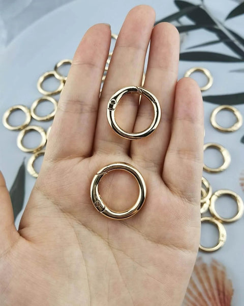 Hand holding two gold 1 inch metal spring O rings with round carabiner clips from 50 Pack 1 Inch Metal Spring O Rings Set With Round Carabiner Clips Snap Hooks For Keychains Purses Shoulder Bags And DIY Crafts In Gold Finish Durable And Handy For Busy DIYers On The Go.