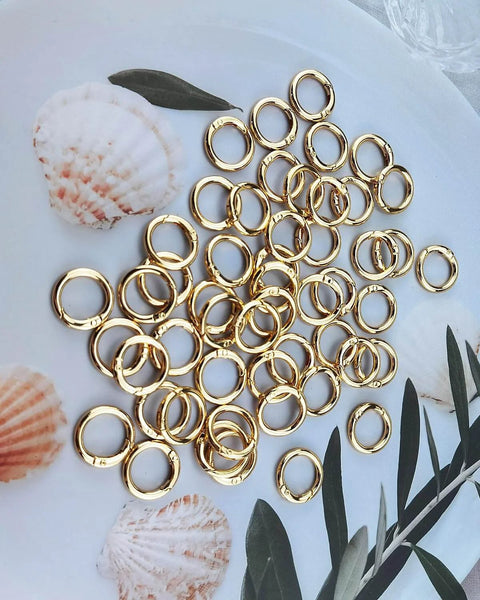 50 Pack 1 Inch Metal Spring O Rings Set With Round Carabiner Clips Snap Hooks For Keychains Purses Shoulder Bags And DIY Crafts In Gold Finish displayed on plate with seashells.