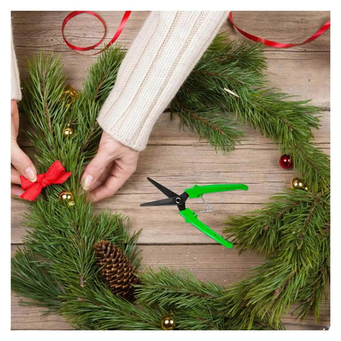 Person crafting holiday wreath with green-handled garden scissors, pine needles, pinecone, and red ribbon on wooden table.