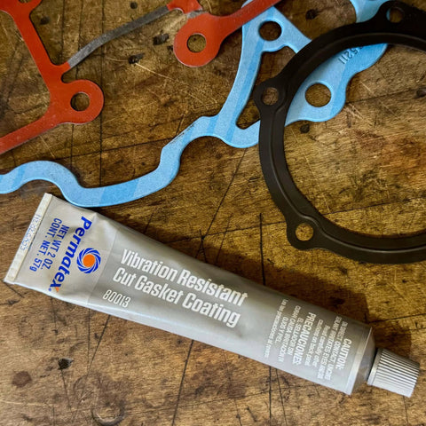 Grey Cut Gasket Coating Sealant For Tough Torque Engines, Vibration Resistant Silicone Sealant For Paper Cork And Rubber Gaskets, Extends Gasket Life And Prevents Blowouts, Fills Scars, Tack Free In 1 Hour, 2 Oz tube on wooden background, with various colored gaskets beside it.