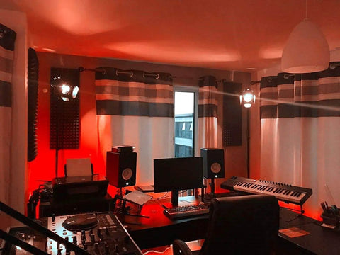 Music studio with red lighting, featuring a desk with monitor, keyboard, and sound equipment, surrounded by soundproofing panels.