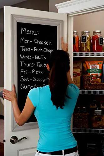 Person adjusting a blackboard menu on a pantry door, showing weekly meal plan and stocked shelves with food items.