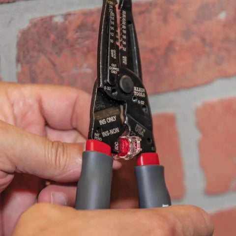 Close-up of hands using Kurve Wire Stripper Crimper Cutter Multi Tool for 10-26 AWG solid and 12-28 AWG stranded wire against a brick wall