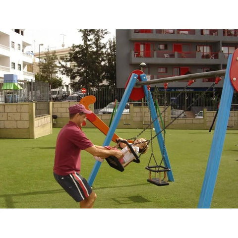 Adult pushing a child on a blue and red swing set in a playground with grass and buildings in the background.