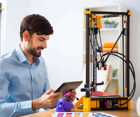 Man operating a 3D printer with a tablet, surrounded by printed objects and color samples on a desk in an office setting.