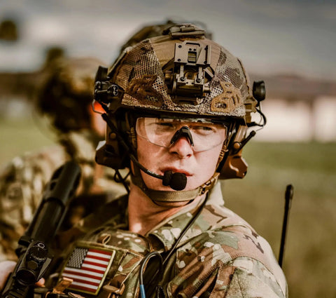 Soldier in camouflage helmet with matte black frames shooting glasses, ANSI Z87 safety and changeable lenses in outdoor setting.