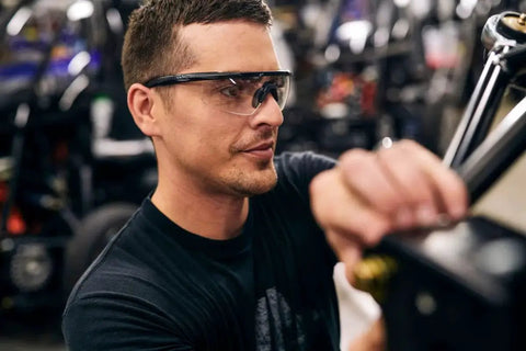 Man wearing Matte Black Frames Shooting Glasses With ANSI Z87 Safety And Changeable Lenses, focused on task in workshop.