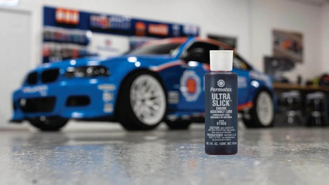 Close-up of Permatex 81950 Ultra Slick Engine Assembly Lube, 4 oz., Red bottle with a blue race car in the background.