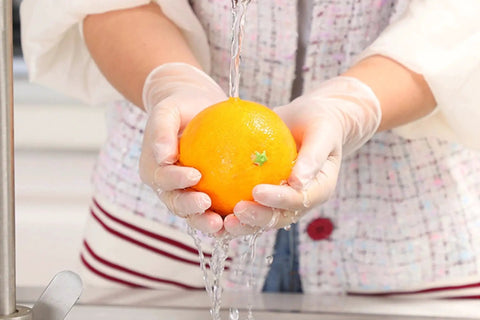 Person wearing Powder Free Vinyl Gloves 4.3 Mil Clear Latex Free For Food Service Cleaning, washing an orange under tap water.