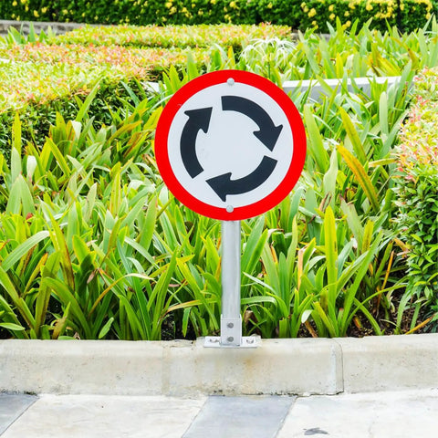Roundabout sign on metal pole in front of green hedge and plants