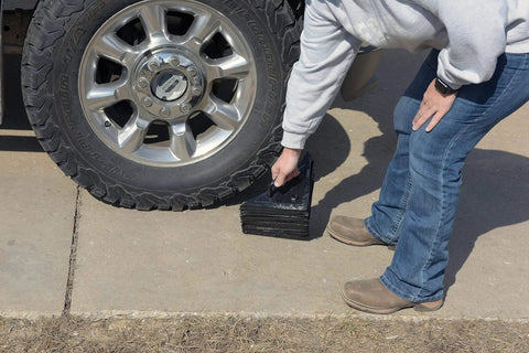 Person installing Durable Laminated Rubber Wheel Chock With Sturdy Metal Handle For Quick Safe Vehicle Blocking On Wet Slippery Or Uneven Surfaces Heavy Duty And Weather Resistant Black Vestil LWC 15 under truck tire