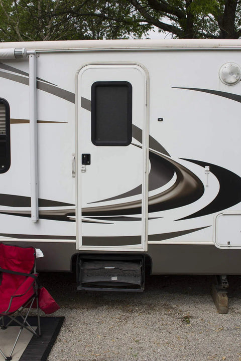 Exterior view of a recreational vehicle with a white door and abstract swirl design. A red chair sits nearby on gravel.