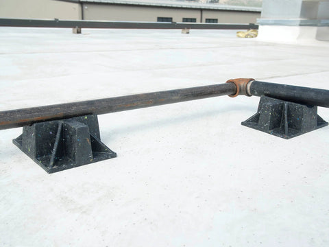Saddle rooftop pipe stand with two black plastic cradles supporting a metal pipe on a concrete roof.