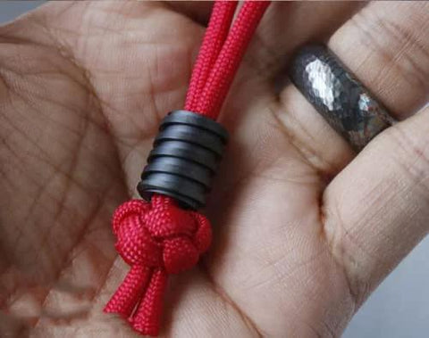 Close-up of red parachute cord with black Ti Titanium Screw Grain Paracord Cord Tool Lanyard Beads Charms EDC Pendant in hand.