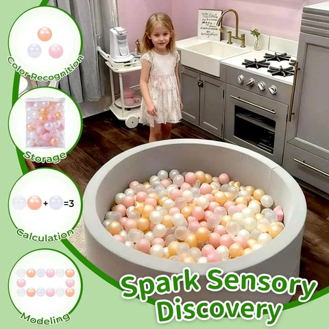 Toddler with gray ball pit filled with plastic balls in pink, gold, pearl white, and clear colors in a kitchen setting.