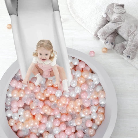 Child sliding into ball pit filled with 100-pack hollow plastic balls in pink, gold, pearl white, clear; safe playtime for toddlers.