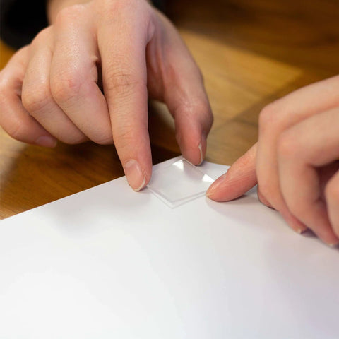 Hands applying Tough And Clear Double Sided Tape Squares For Mounting, 24 Pre-Cut 1-Inch Squares, to a white surface.