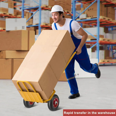 Worker in warehouse using a yellow hand truck with large boxes, wearing blue overalls and white hard hat.