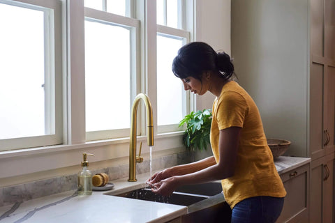 Woman washing hands at a kitchen sink with gold faucet and marble countertop, potted plant in background