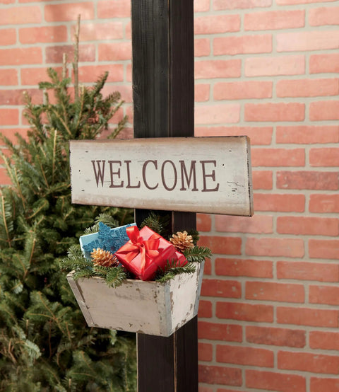Welcome sign with a rustic planter and red gift on a post against a brick wall background with greenery.
